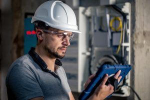 Man, an electrical technician working in a switchboard with fuses. Installation and connection of electrical equipment. Professional uses a tablet. electricien qualifelec Lyon Pro Garantie Assurance Conseil expert certifié